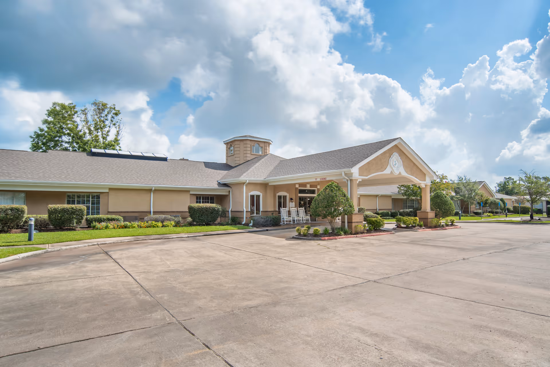 Exterior view of Brookdale Lake Charles senior living facility showing a single-story building with a covered entrance, rocking chairs on the porch, neatly trimmed bushes, and a large paved driveway under a partly cloudy sky.