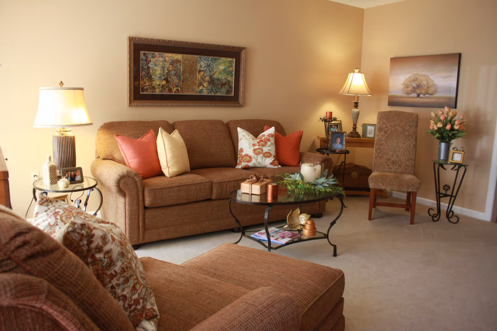 A cozy living room with a brown sofa and matching armchair, decorated with colorful throw pillows. There is a glass coffee table with decorative items and magazines underneath. Two side tables hold lamps, framed photos, and a vase with flowers. The walls are beige with framed artwork hanging.