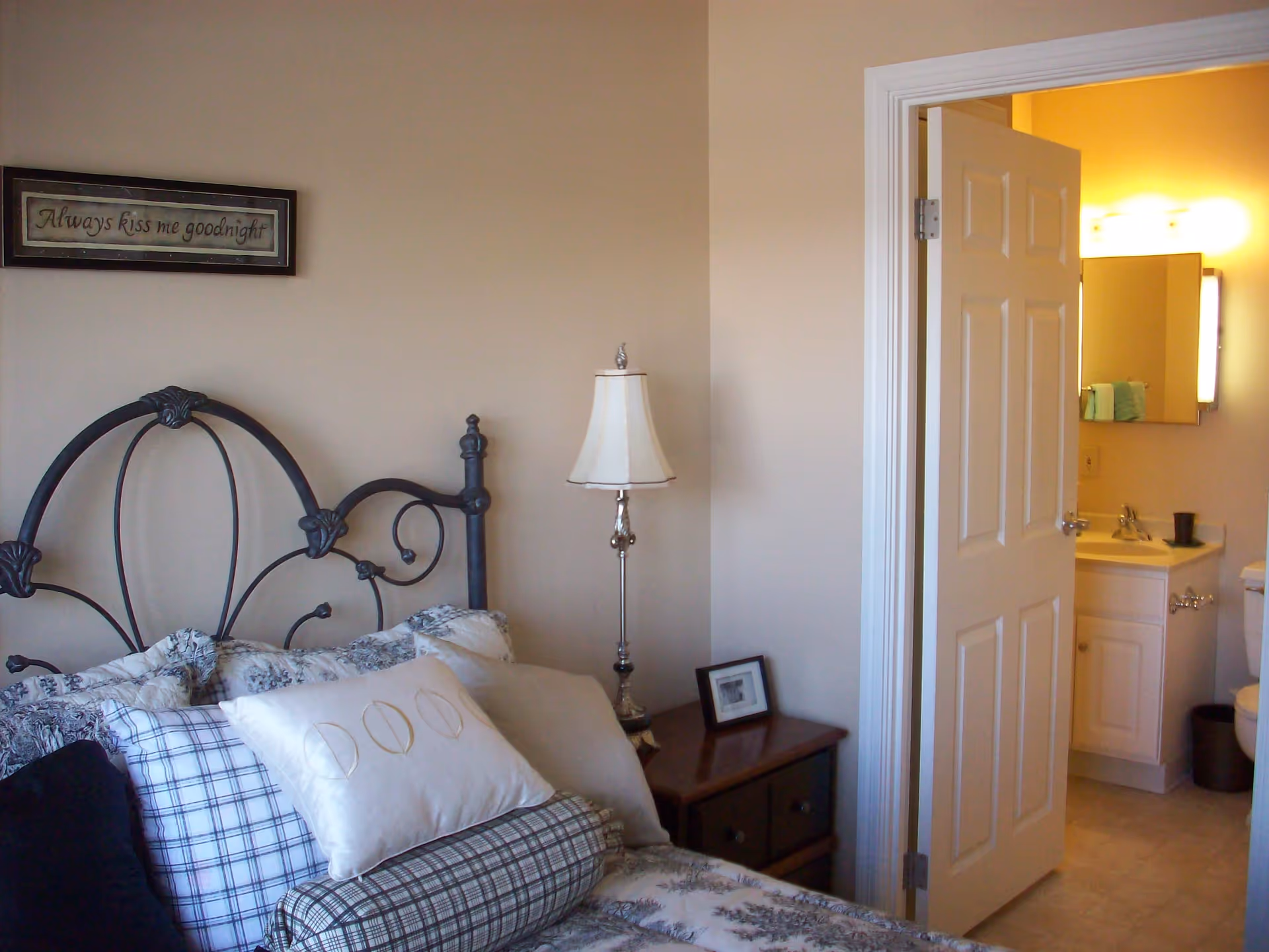 A bedroom with a decorative metal headboard, multiple pillows on the bed, a bedside table with a lamp and a small framed picture. The door to an adjoining bathroom is open, showing a sink, mirror, and toilet inside.