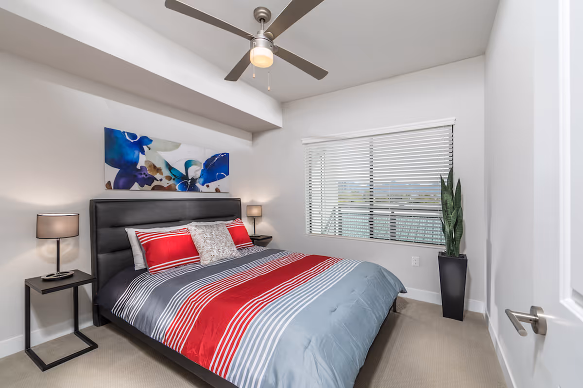 Modern bedroom with a bed dressed in red, gray, and white striped bedding, bedside tables and lamps, a ceiling fan, window blinds, and a potted plant.
