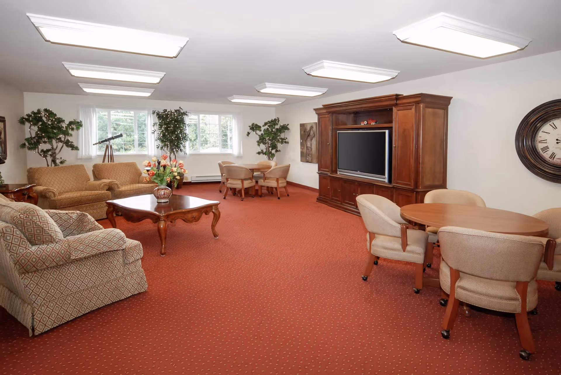 A spacious living room with red carpet, beige upholstered chairs and sofas, a wooden coffee table with a flower vase, a large wooden entertainment center with a TV, round tables with chairs on wheels, large windows with white curtains, and decorative plants and wall clock.
