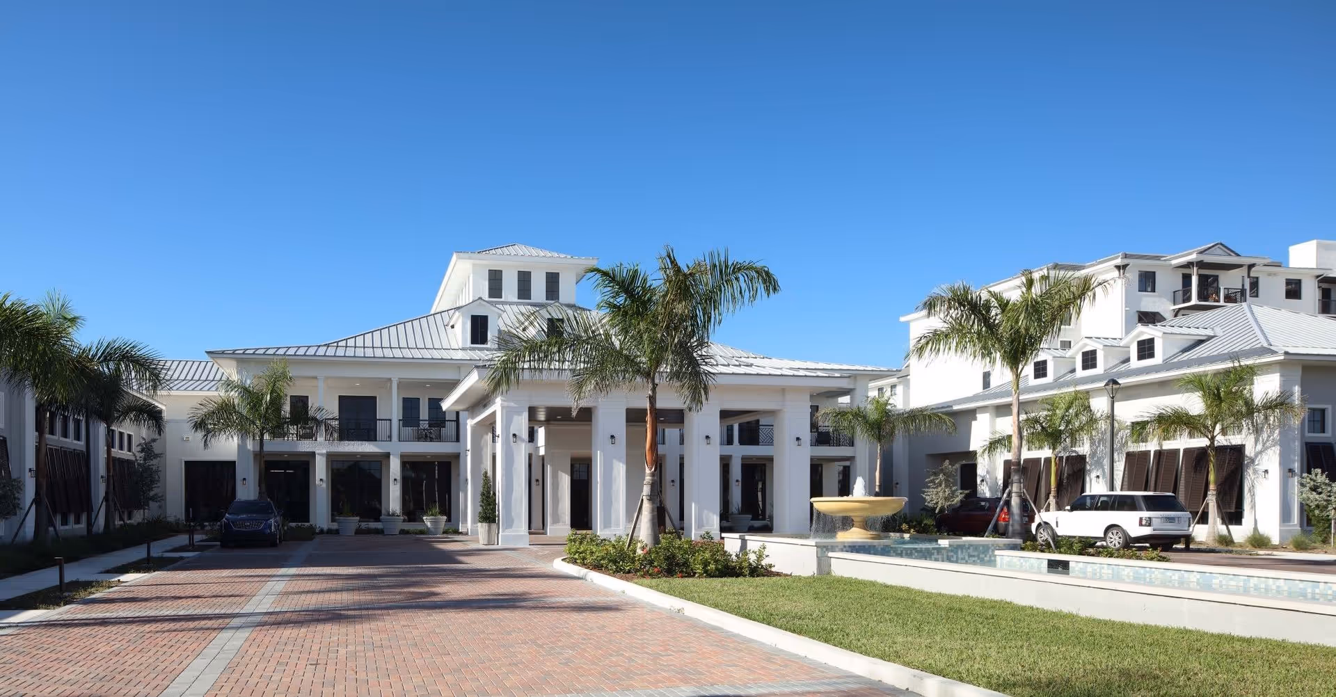 Front entrance of a white coastal-style senior living building with palm trees, a fountain, and a brick driveway under a clear blue sky.