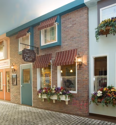 Indoor scene resembling a quaint street with brick and pastel-colored walls, featuring a cafe storefront with green door, striped red awnings, flower boxes with colorful flowers, and warm wall-mounted lanterns.