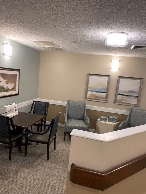 A small seating area in a senior living facility with a dark wooden table surrounded by four chairs, two upholstered armchairs, a small side table with two decorative boxes, and framed artwork on the walls. The room has soft lighting from wall sconces and a ceiling light.