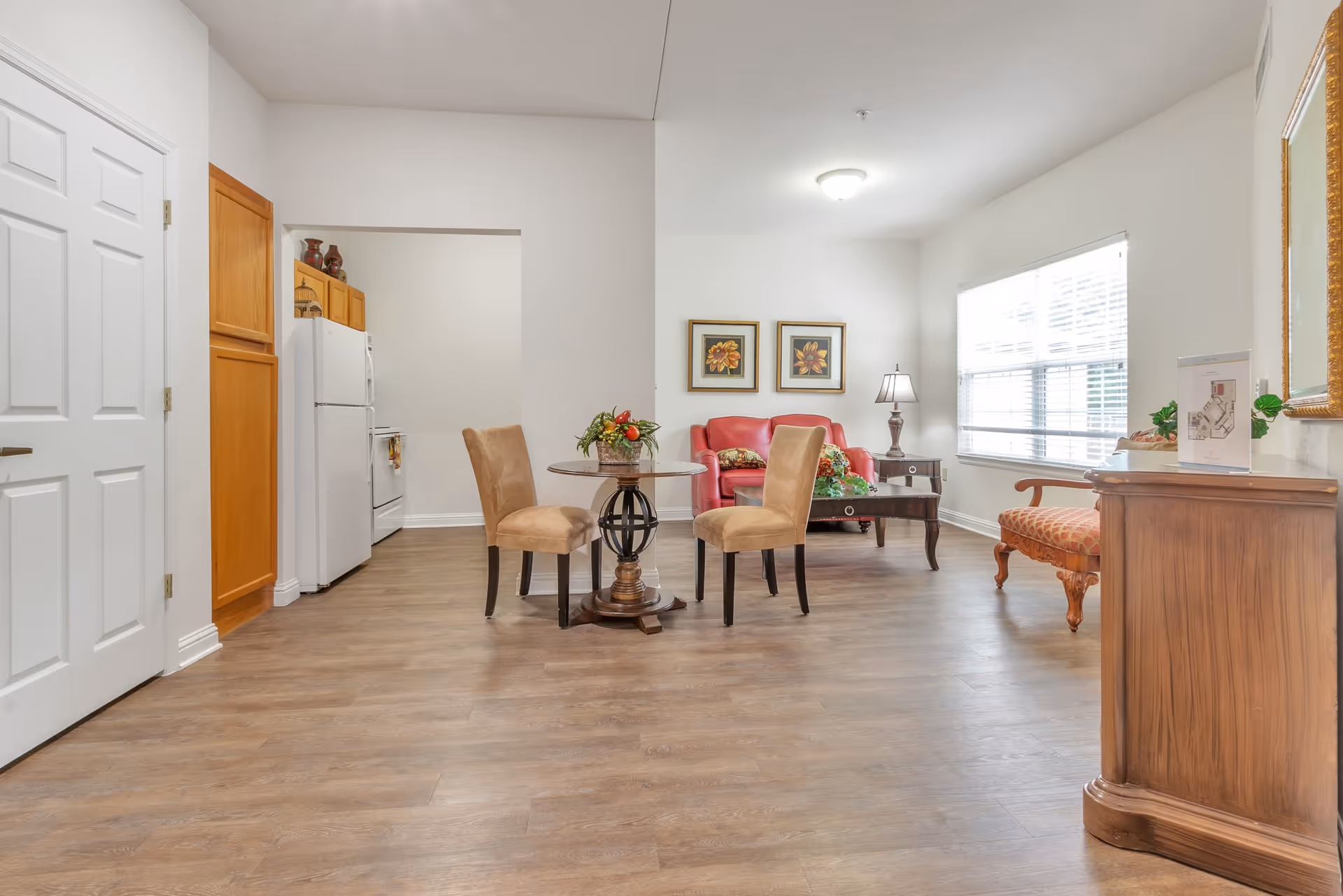 A bright and spacious interior room featuring a small round dining table with two beige chairs, a red loveseat with floral cushions, a wooden coffee table, a side table with a lamp, a large window with blinds, and a kitchen area with white appliances and wooden cabinets in the background.