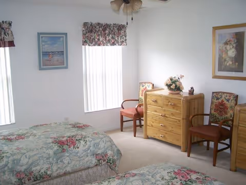 Bright bedroom with two floral twin beds, a wooden dresser, two chairs, framed artwork, and vertical blinds.