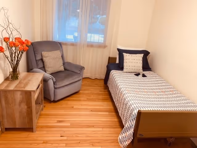 A small, cozy bedroom with a single bed covered in a patterned quilt and two pillows, a gray cushioned armchair with a striped pillow, a wooden nightstand with a vase of orange flowers, and a window with sheer white curtains letting in natural light.