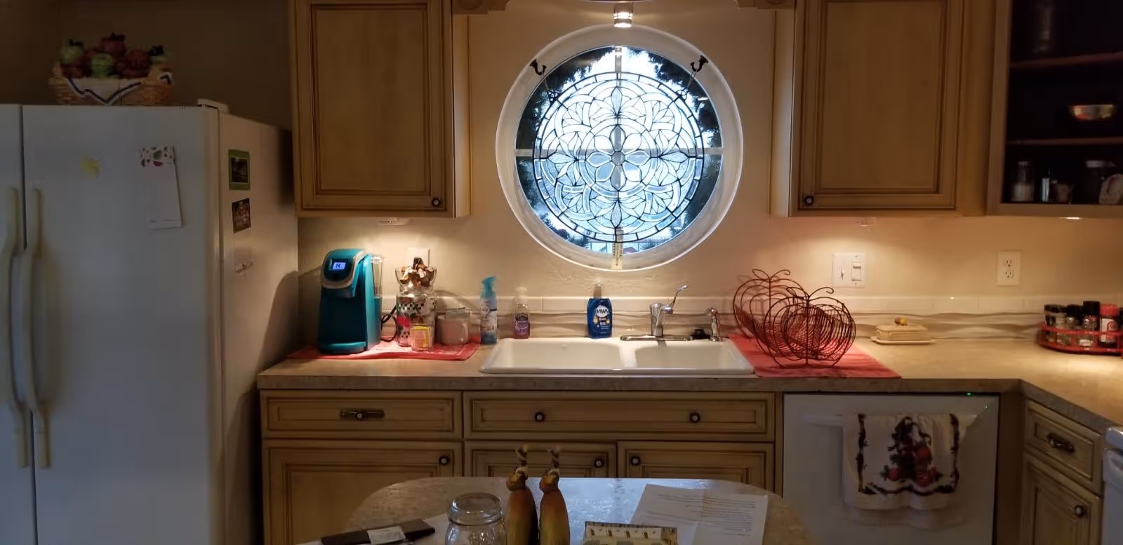 A kitchen interior featuring a white refrigerator on the left, beige cabinets, and a countertop with a teal coffee maker, various bottles, and a double sink beneath a round window with decorative stained glass. A kitchen island with papers and salt and pepper shakers is in the foreground.