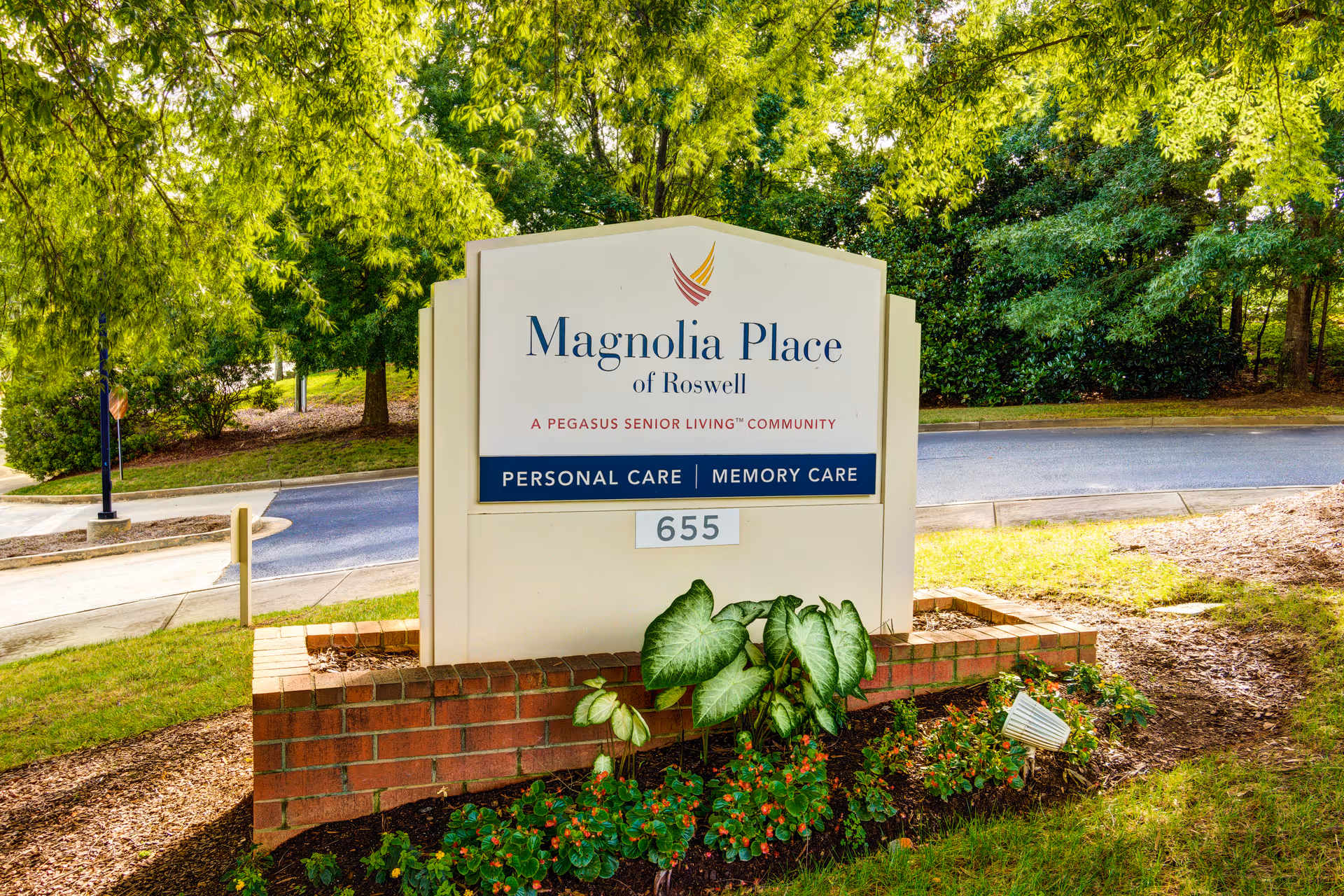 Outdoor image of a sign for Magnolia Place of Roswell, a Pegasus Senior Living community offering personal care and memory care services. The sign is surrounded by greenery, plants, and trees with a road in the background.