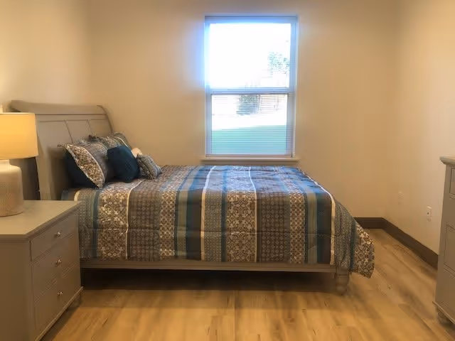 A neatly made bed with a patterned blue and gray bedspread and multiple pillows in a bright bedroom with light-colored walls and wooden flooring. There is a window above the bed letting in natural light, a nightstand with a lamp on the left side, and a dresser on the right side.
