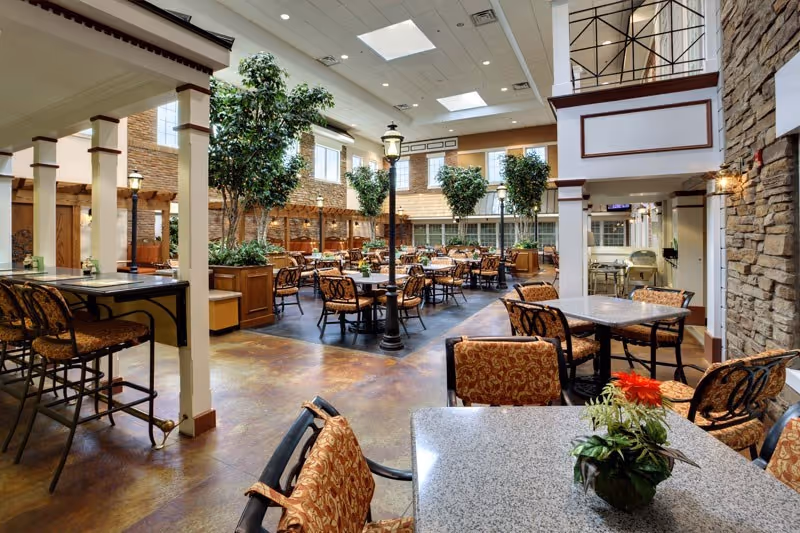 Spacious dining area with multiple tables and chairs arranged neatly, decorated with potted plants and indoor trees. The room features high ceilings with skylights, stone accent walls, and warm lighting, creating a welcoming atmosphere.