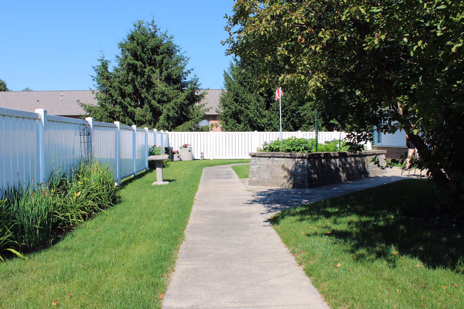 Sunlit courtyard with a concrete walkway flanked by a white fence, raised garden bed, birdbath and trees.