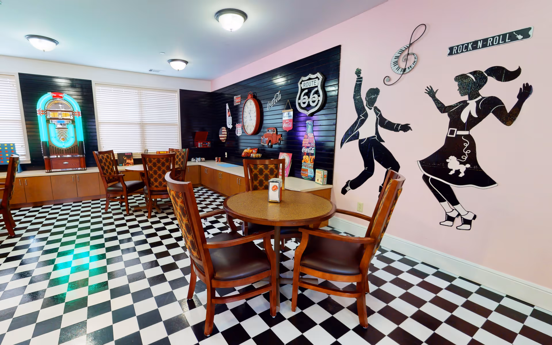 A retro-themed dining area with black and white checkered floor tiles, wooden tables and chairs, a jukebox near the windows, and wall decorations including a Route 66 sign, a large clock, and silhouettes of a dancing couple with a 'Rock-N-Roll' sign on a pink wall.