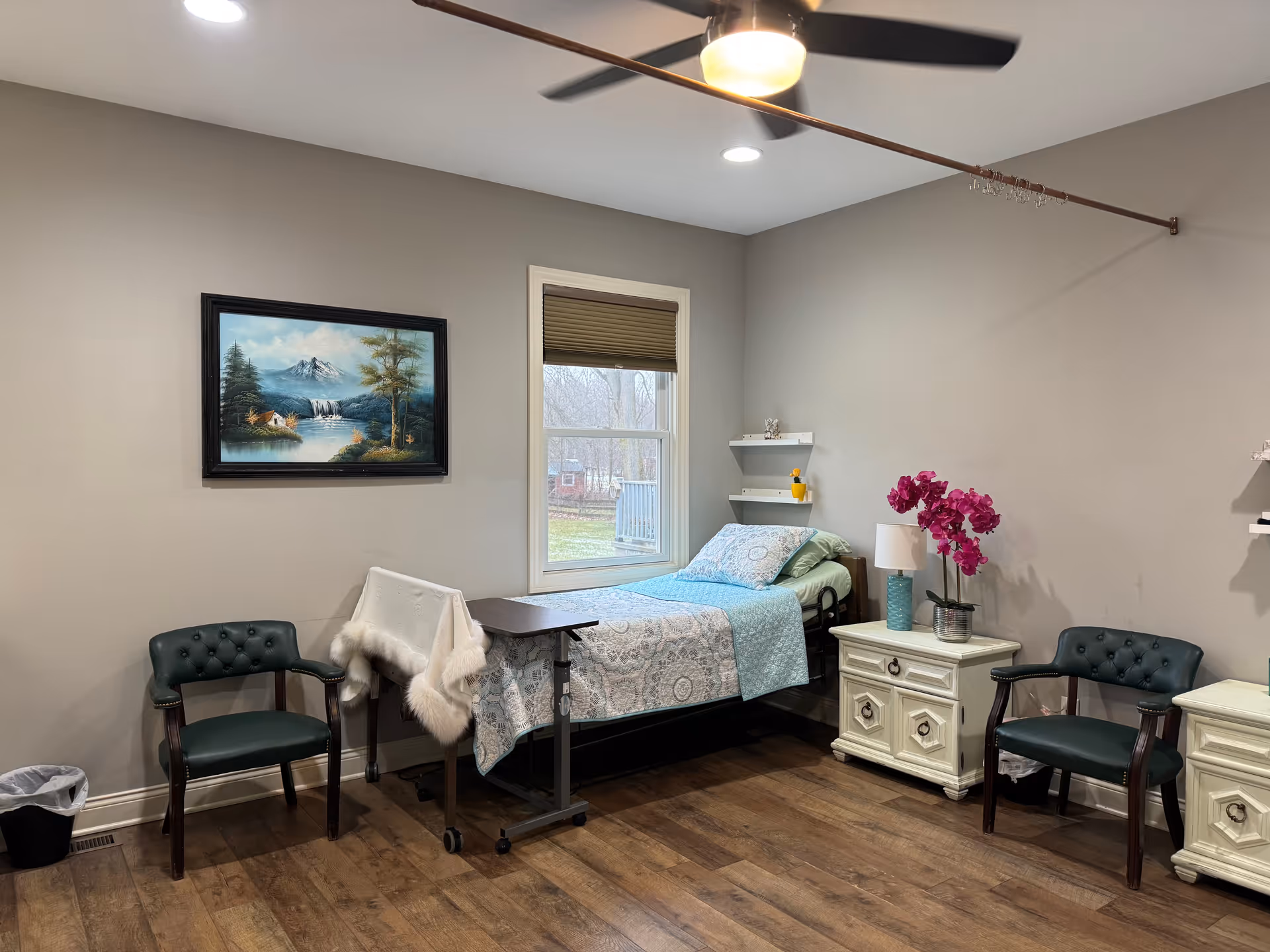 A neatly arranged senior bedroom with a single bed covered in a patterned blue and white quilt. The room has two dark green cushioned chairs, two white nightstands with drawers, a lamp, and a vase with pink flowers. A window with a beige blind lets in natural light, and a ceiling fan with a light is mounted above. A landscape painting hangs on the wall, and the floor is wooden.