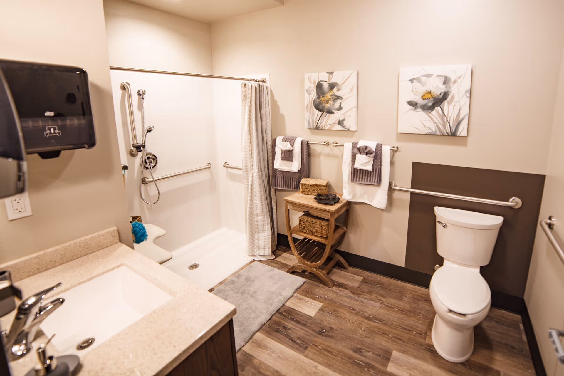 A clean and accessible bathroom featuring a walk-in shower with grab bars and a shower seat, a toilet with grab bars on both sides, a sink with a countertop, a paper towel dispenser, two floral paintings on the wall, a small wooden table with baskets, and neatly folded towels hanging on a towel rack.