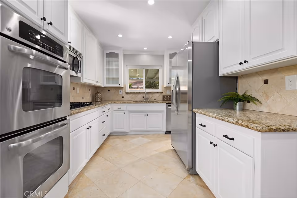 A modern kitchen with white cabinets, granite countertops, and stainless steel appliances including a double oven, microwave, refrigerator, and dishwasher. The kitchen has beige tiled flooring and a window above the sink allowing natural light to enter.