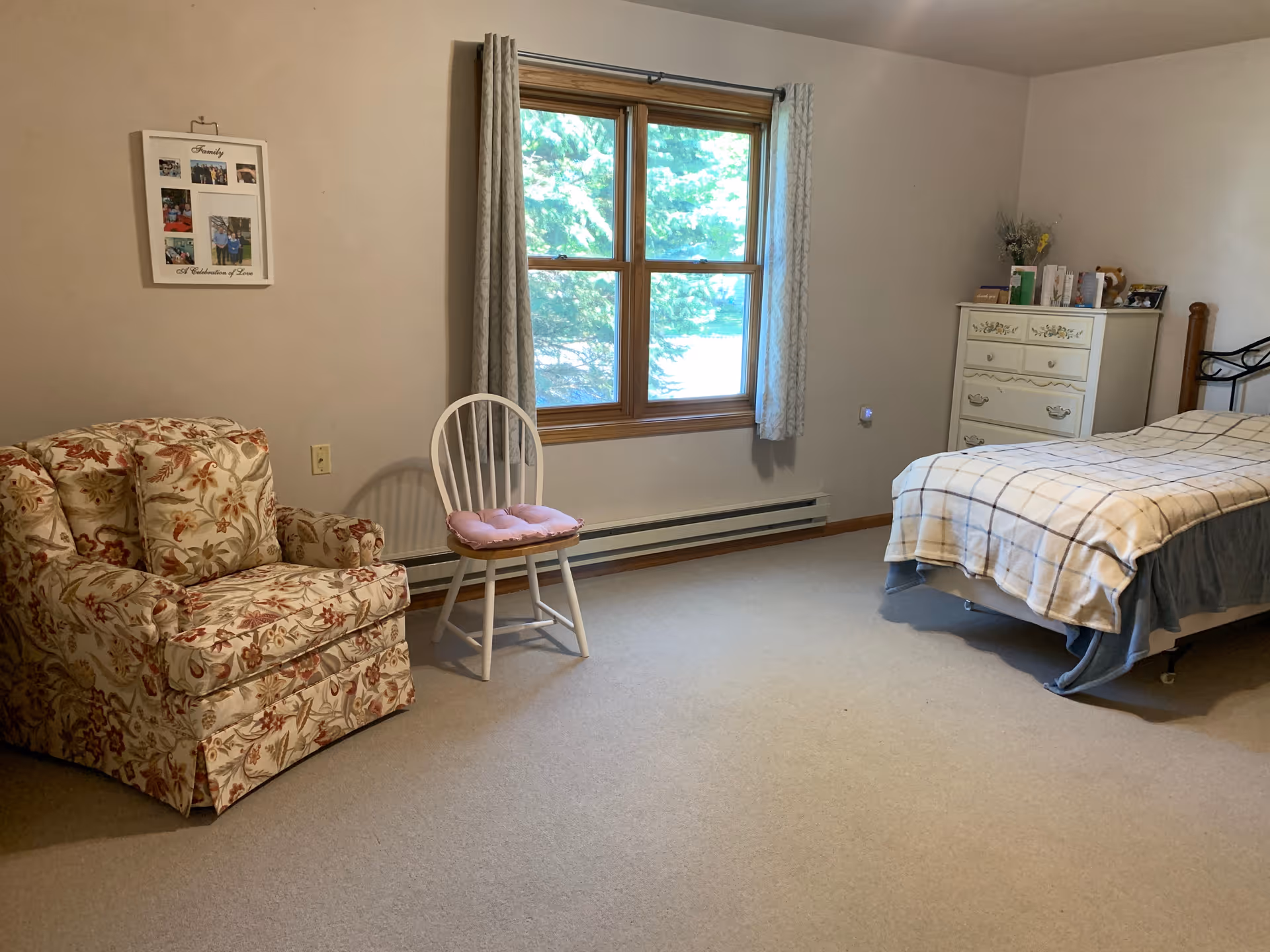 A cozy bedroom with a floral armchair and a white wooden chair with a pink cushion near a window with light gray curtains. There is a bed with a checkered blanket and a white dresser with decorative items on top. The walls are light-colored and the floor is carpeted.