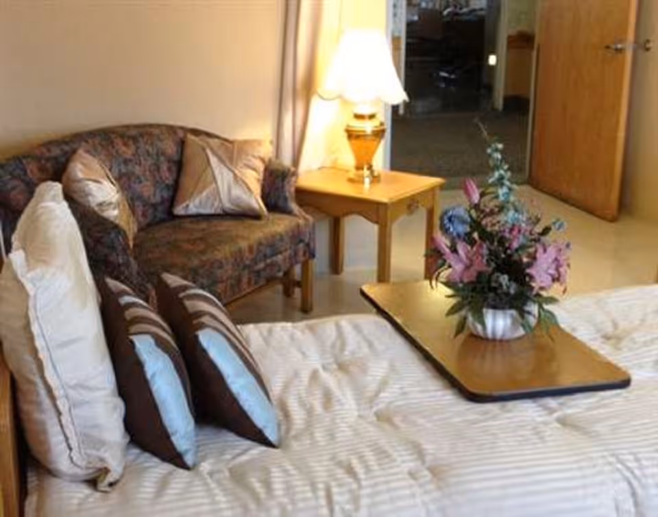 A cozy bedroom area featuring a bed with multiple pillows and a wooden tray holding a vase of colorful flowers. Next to the bed is a small wooden side table with a lit lamp. In the background, there is a patterned loveseat with decorative pillows and an open door leading to another room.