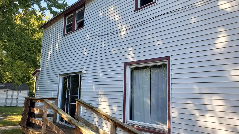 Two-story white vinyl-sided house exterior with a sliding glass door, a curtained window, and a wooden ramp with railings leading to the entrance, set among trees.