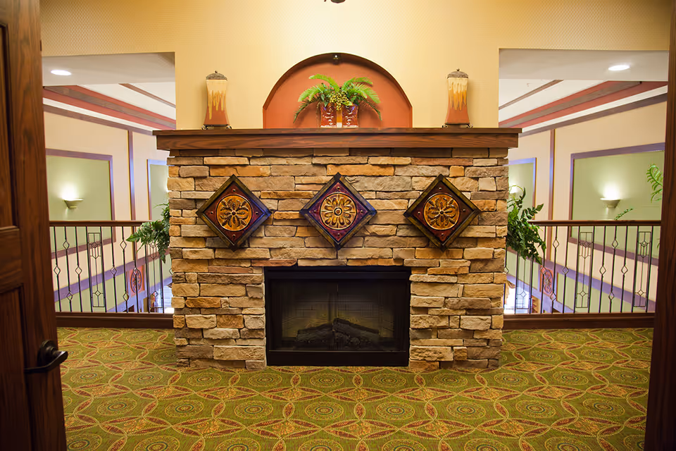 Interior view of a senior living facility featuring a stone fireplace with three decorative square plaques mounted above it. The fireplace mantel holds two candle holders and a potted plant. The area has patterned green carpet and is flanked by railings overlooking a lower level with wall sconces and decorative trim.