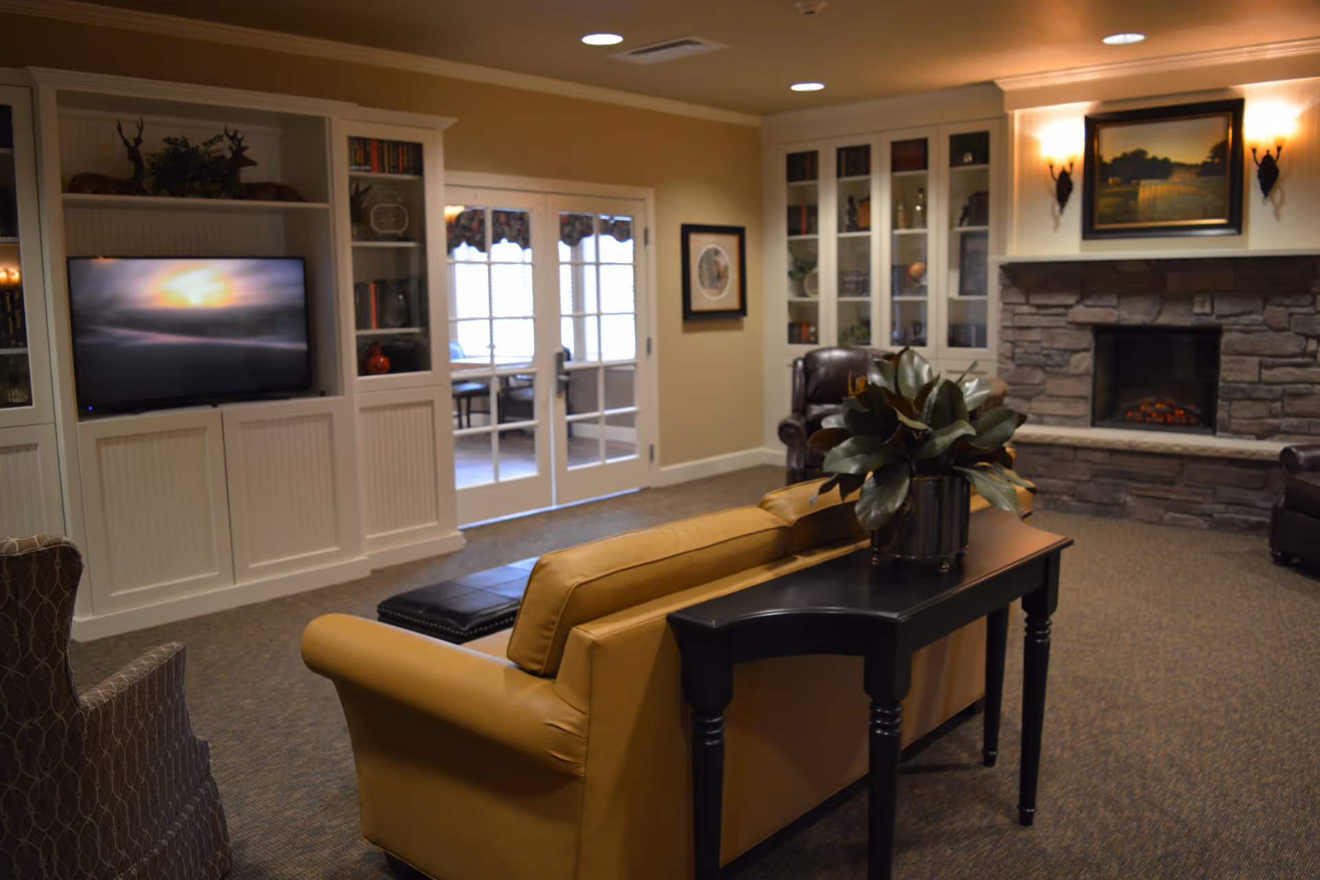 A cozy living room area in Edgemont Place featuring a tan leather sofa, a black side table with a potted plant, a stone fireplace with a painting above it, built-in white shelves with books and decorations, a flat-screen TV, and glass double doors leading to another room.