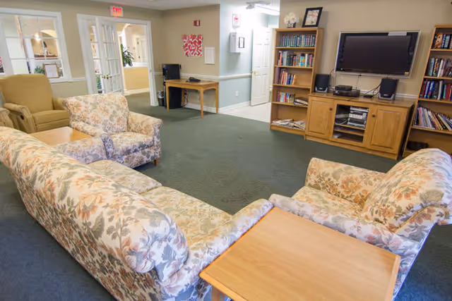 A cozy living room area with floral patterned sofas and armchairs arranged around a wooden coffee table. There is a wooden entertainment center with a flat-screen TV and shelves filled with books. The room has a green carpet and light-colored walls with a small desk and chair in the background near a hallway.