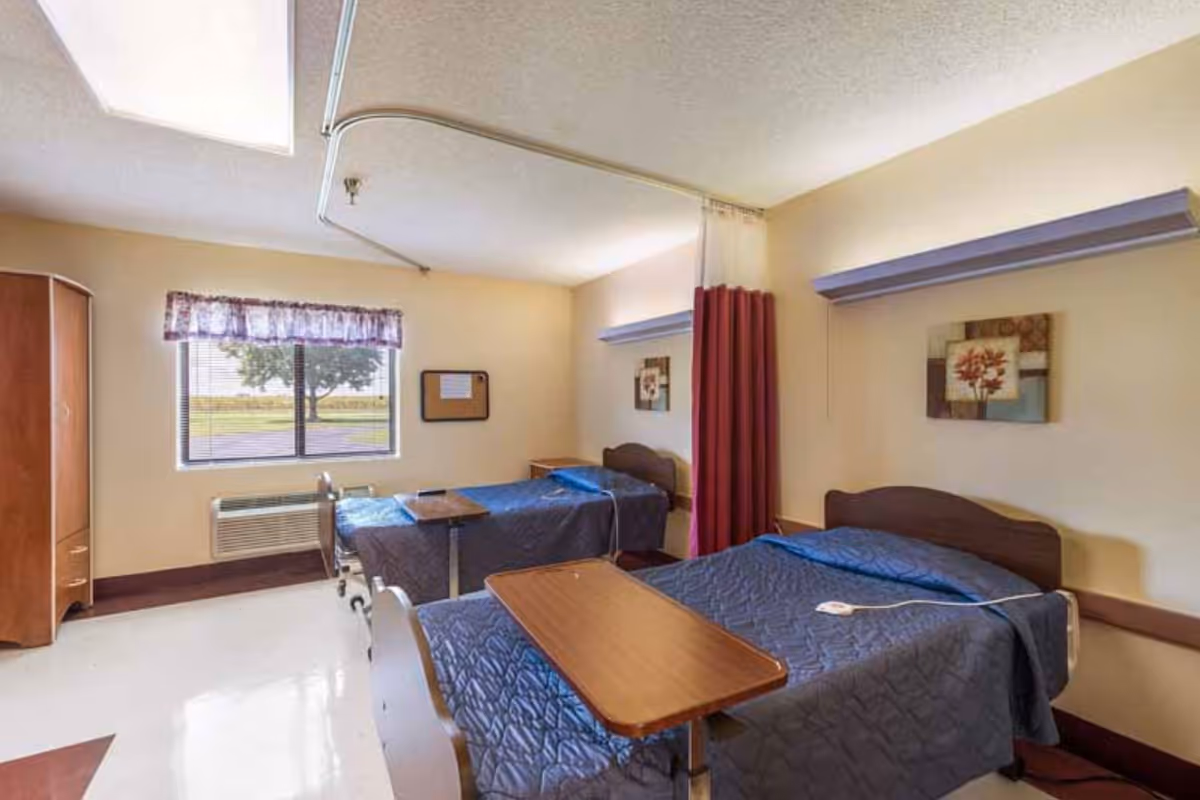 Two beds with blue quilts in a shared bedroom with window and furniture.