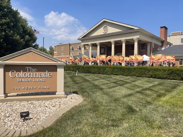 Front exterior of The Colonnade Senior Living with a columned entrance, landscaped lawn, and a sign reading the community name and address.