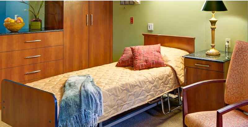 A single bed with a beige quilted bedspread and two red patterned pillows in a room with wooden cabinets and a green wall. A blue-gray throw blanket is draped over the foot of the bed. Next to the bed is a wooden nightstand with a black granite top and a brass lamp. An upholstered chair with a red patterned fabric is partially visible in the foreground.