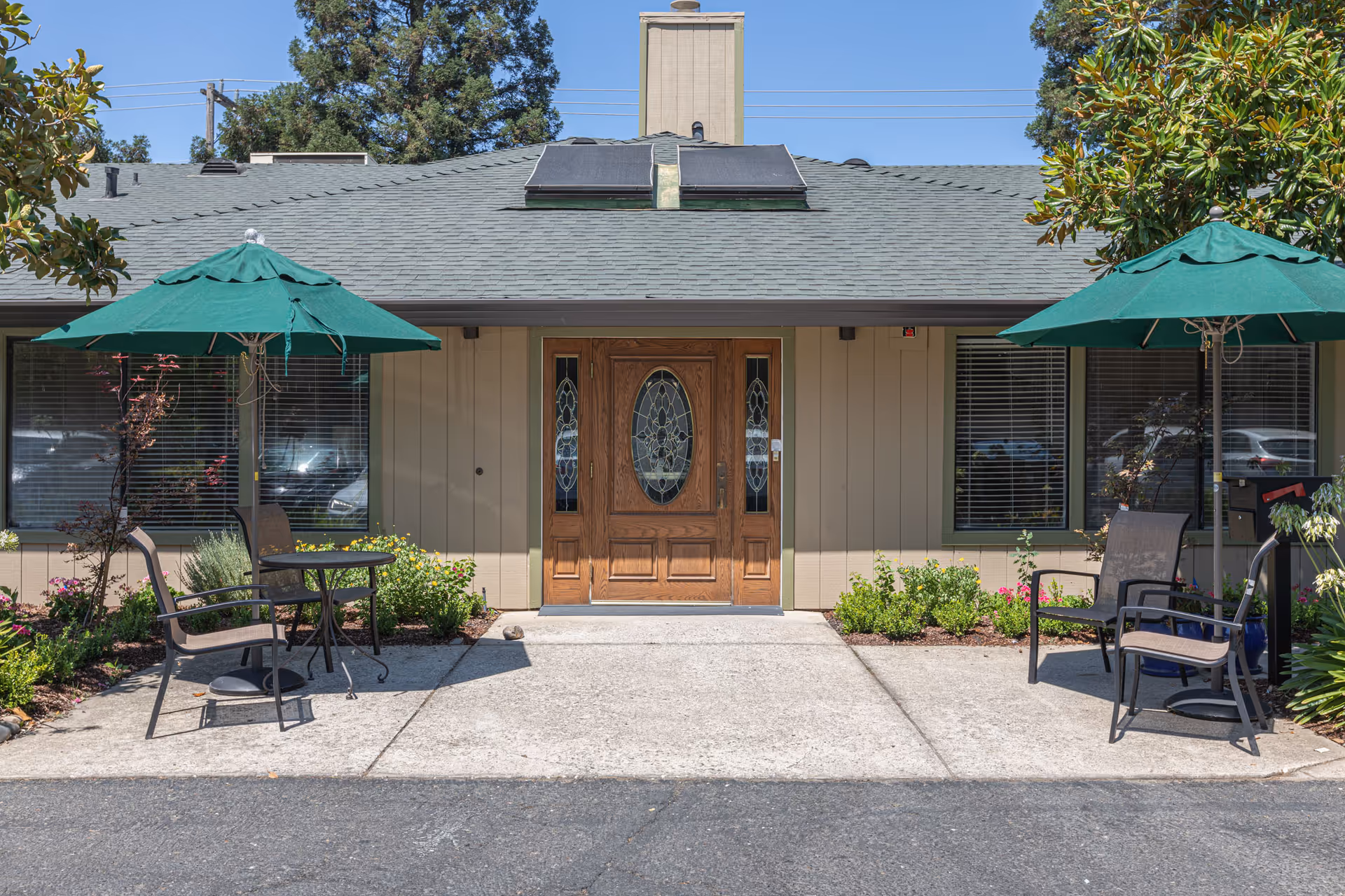 Front entrance of a single-story building with a wooden door featuring decorative glass panels. There are two green patio umbrellas with chairs and small tables on either side of the entrance. The area is landscaped with small bushes and flowers, and there are windows with blinds on both sides of the door.