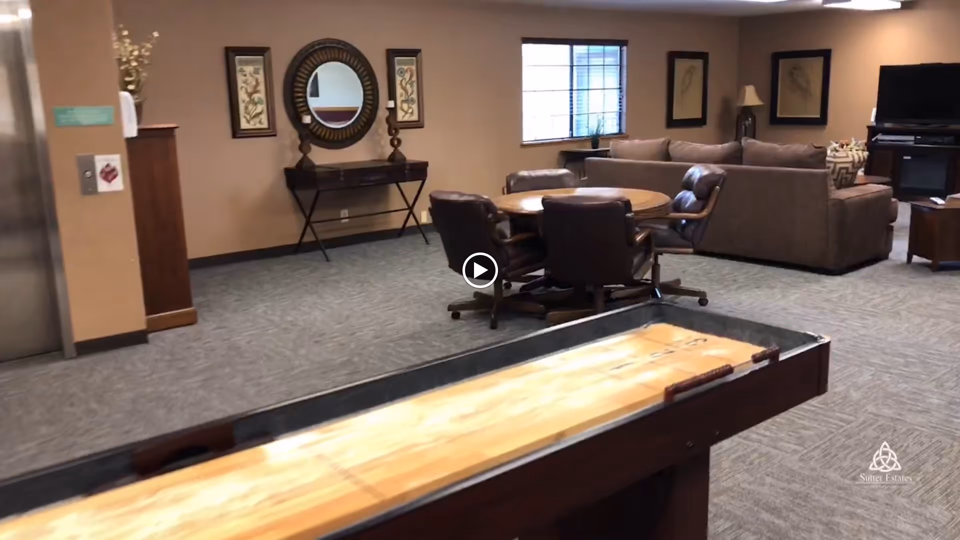A spacious common area in a retirement community featuring a shuffleboard table in the foreground, a round table with four chairs, a large sectional sofa, a TV on a stand, and decorative wall art and a mirror. There is an elevator on the left side of the room.