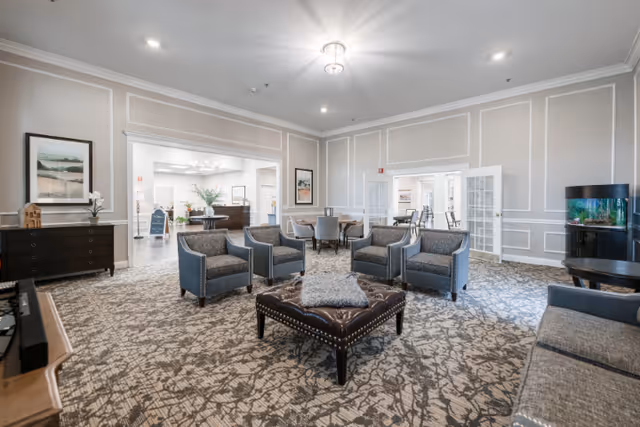 A spacious senior living facility common area with patterned carpet, four gray armchairs arranged around a large tufted leather ottoman, and additional seating including a gray sofa and dining chairs. The room features light gray walls with white trim, framed artwork, a fish tank on a stand, and a view into an adjacent room with more seating and a reception desk.