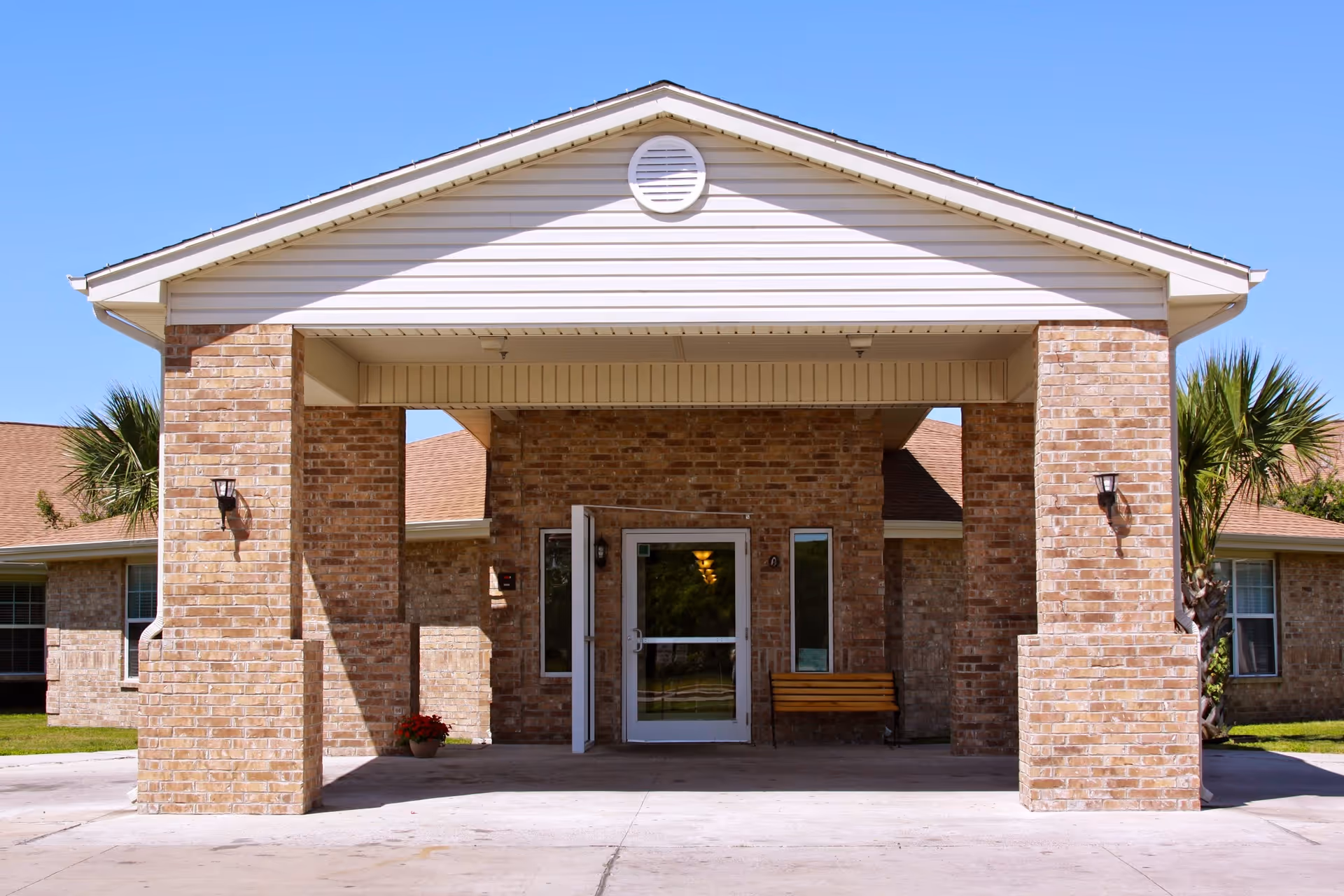 Front entrance of a nursing and rehabilitation center with brick pillars supporting a covered driveway. There is a glass door entrance with windows on either side, a wooden bench to the right, and a potted plant to the left. The building has a light-colored roof and exterior walls made of brick. Palm trees and a clear blue sky are visible in the background.