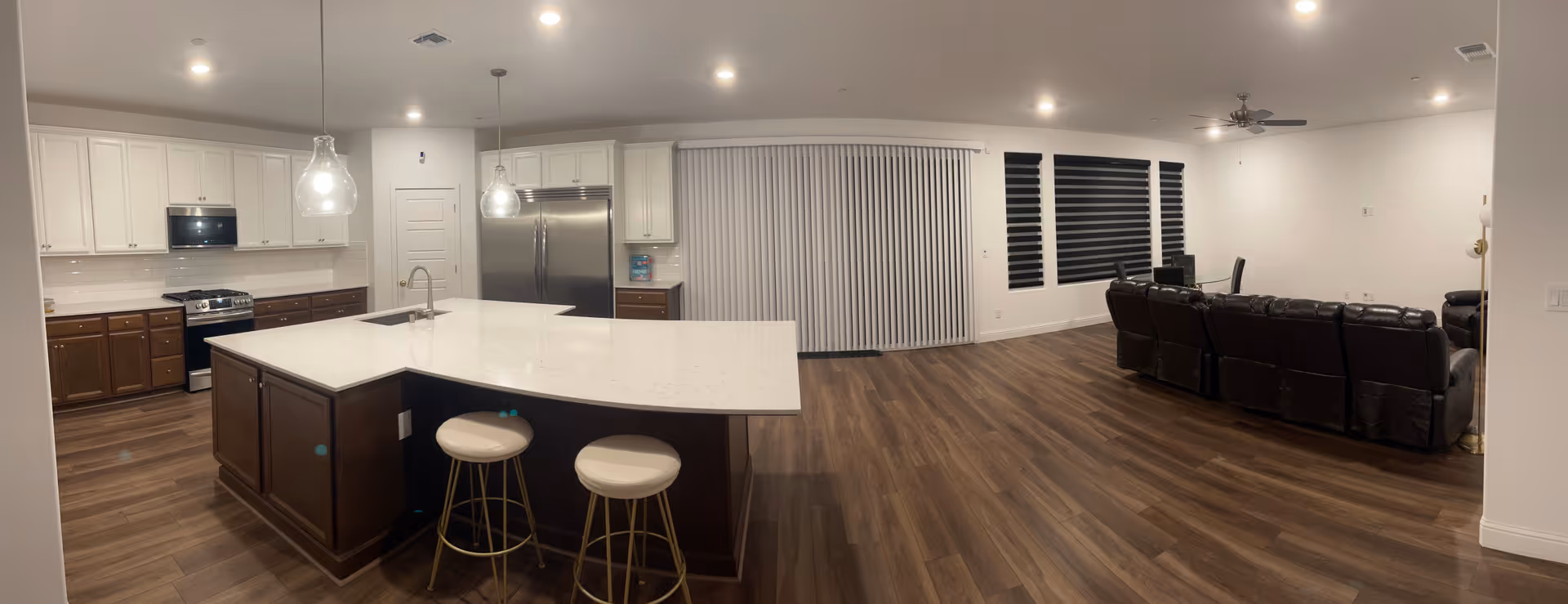 A spacious open-concept interior featuring a modern kitchen with white upper cabinets, dark lower cabinets, a large island with a white countertop, two bar stools, stainless steel appliances, and pendant lighting. Adjacent to the kitchen is a living area with dark leather recliner sofas, wood flooring, large windows with blinds, and a ceiling fan.