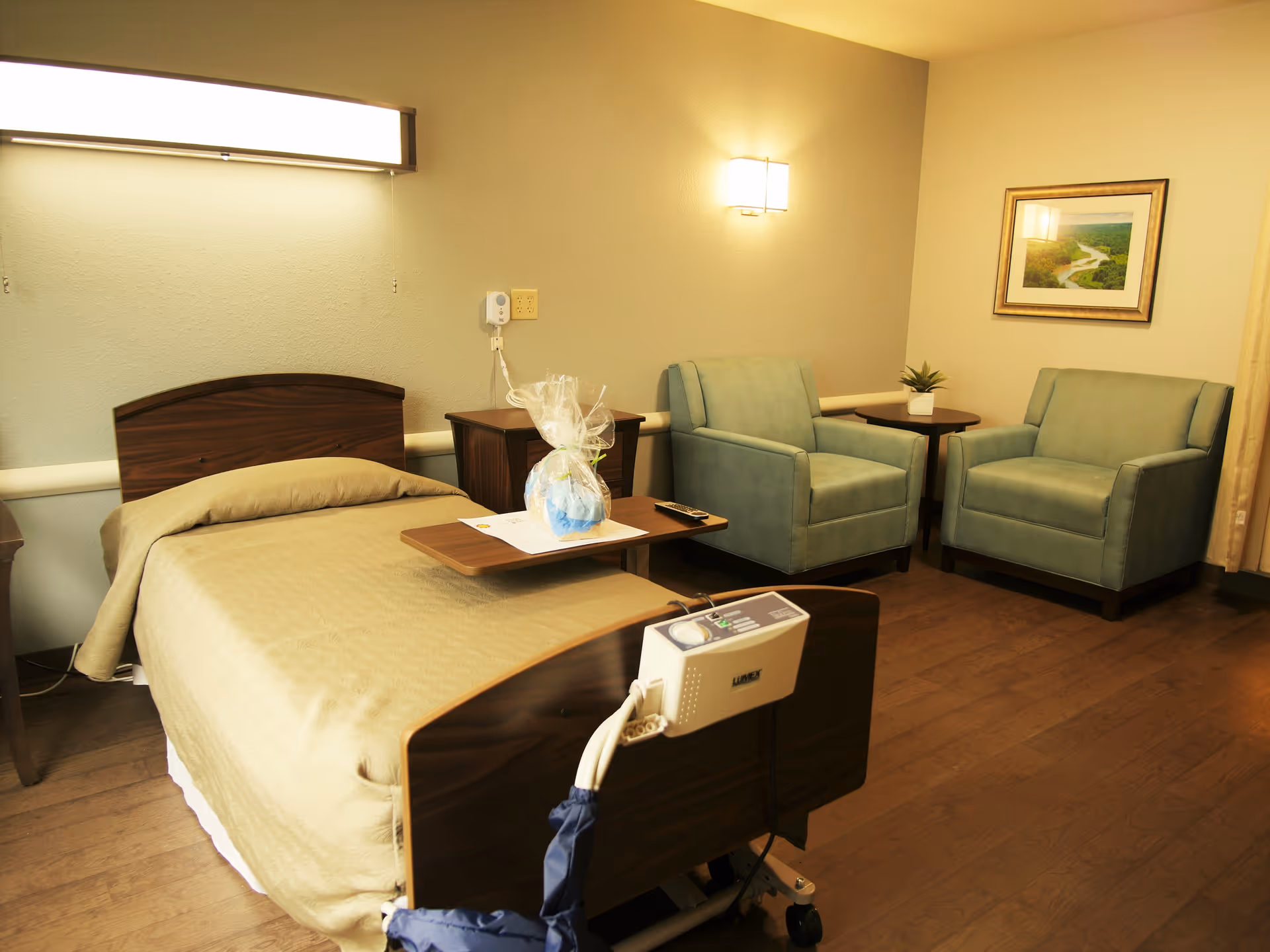 A nursing facility room with a single hospital bed covered with a beige blanket. There is a wooden bedside table with a wrapped gift and a remote control on a tray attached to the bed. Two green upholstered armchairs are positioned near a small wooden table with a potted plant. A framed landscape picture hangs on the wall, and the room has warm lighting with a wall-mounted light fixture.