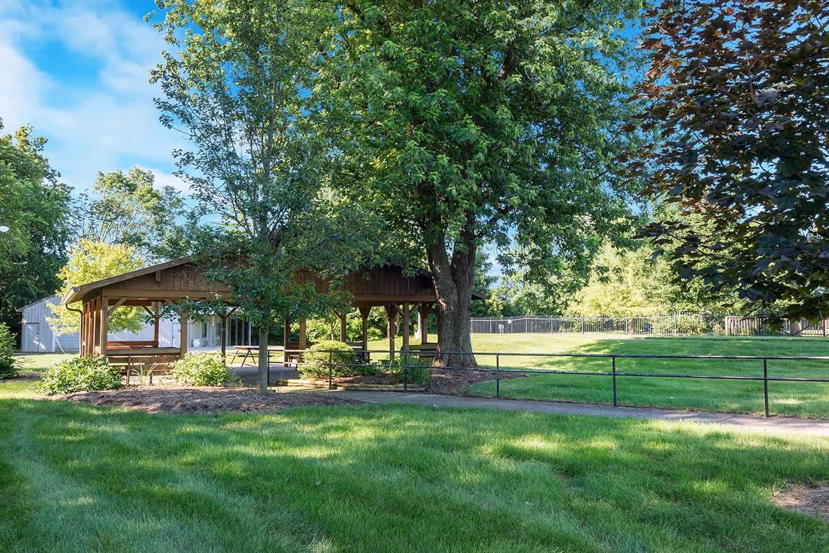 A wooden pavilion with picnic tables underneath surrounded by green grass, trees, and shrubs on a sunny day with a blue sky and some clouds.