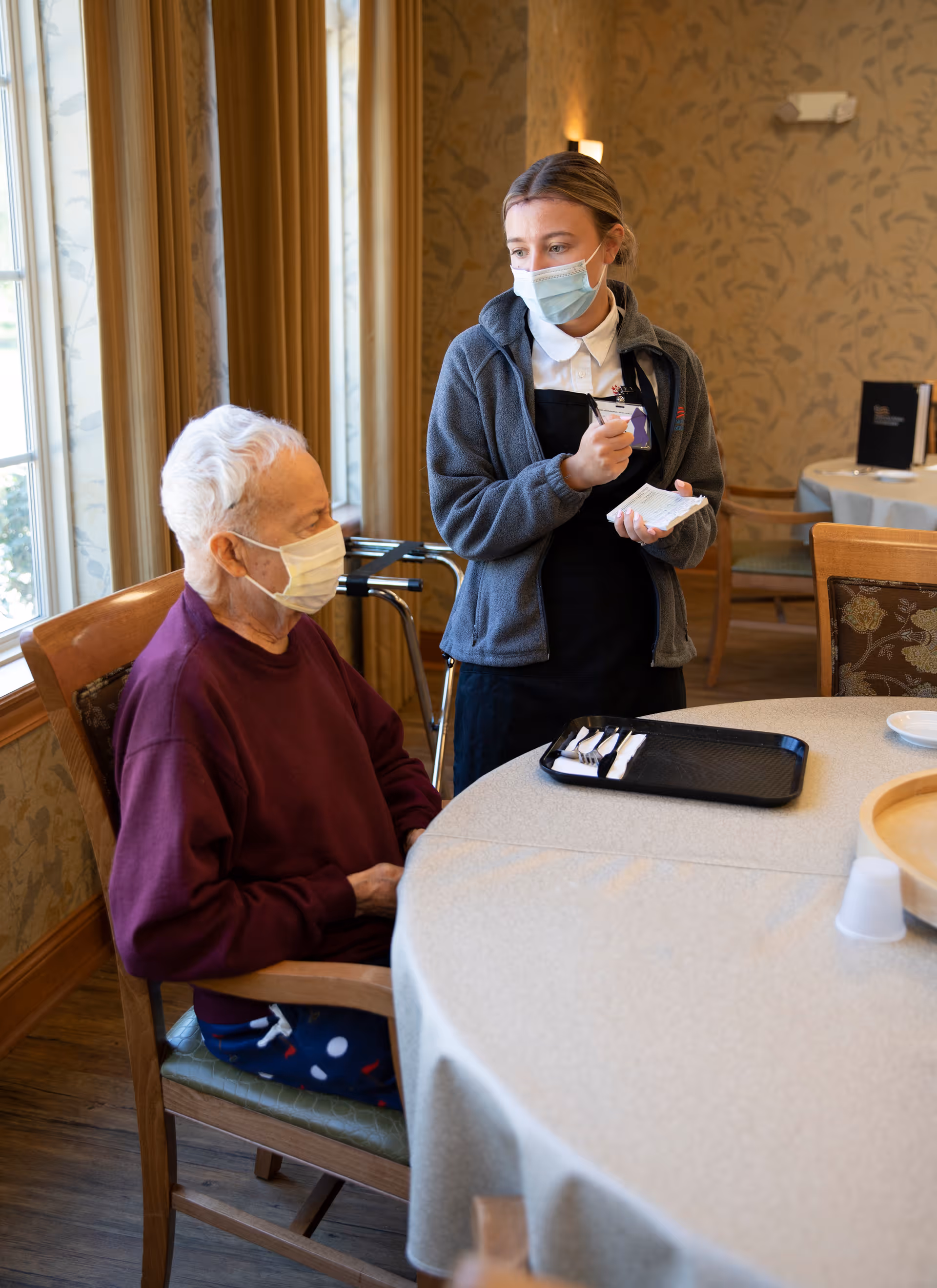 A masked caregiver talks with a masked elderly man seated at a round dining table in a dining room.