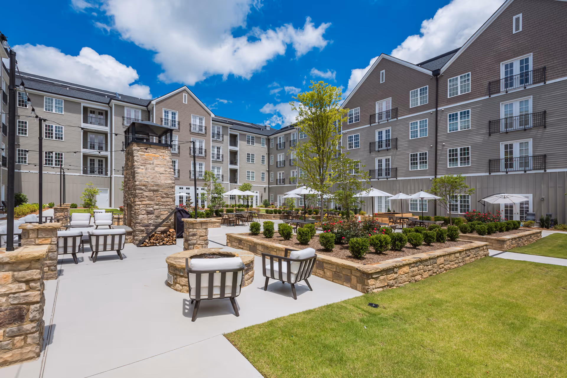 Outdoor courtyard area of a senior living facility with stone walls, patio seating with cushioned chairs, tables with umbrellas, landscaped garden beds, and a tall stone fireplace under a blue sky with scattered clouds. The multi-story building surrounds the courtyard.