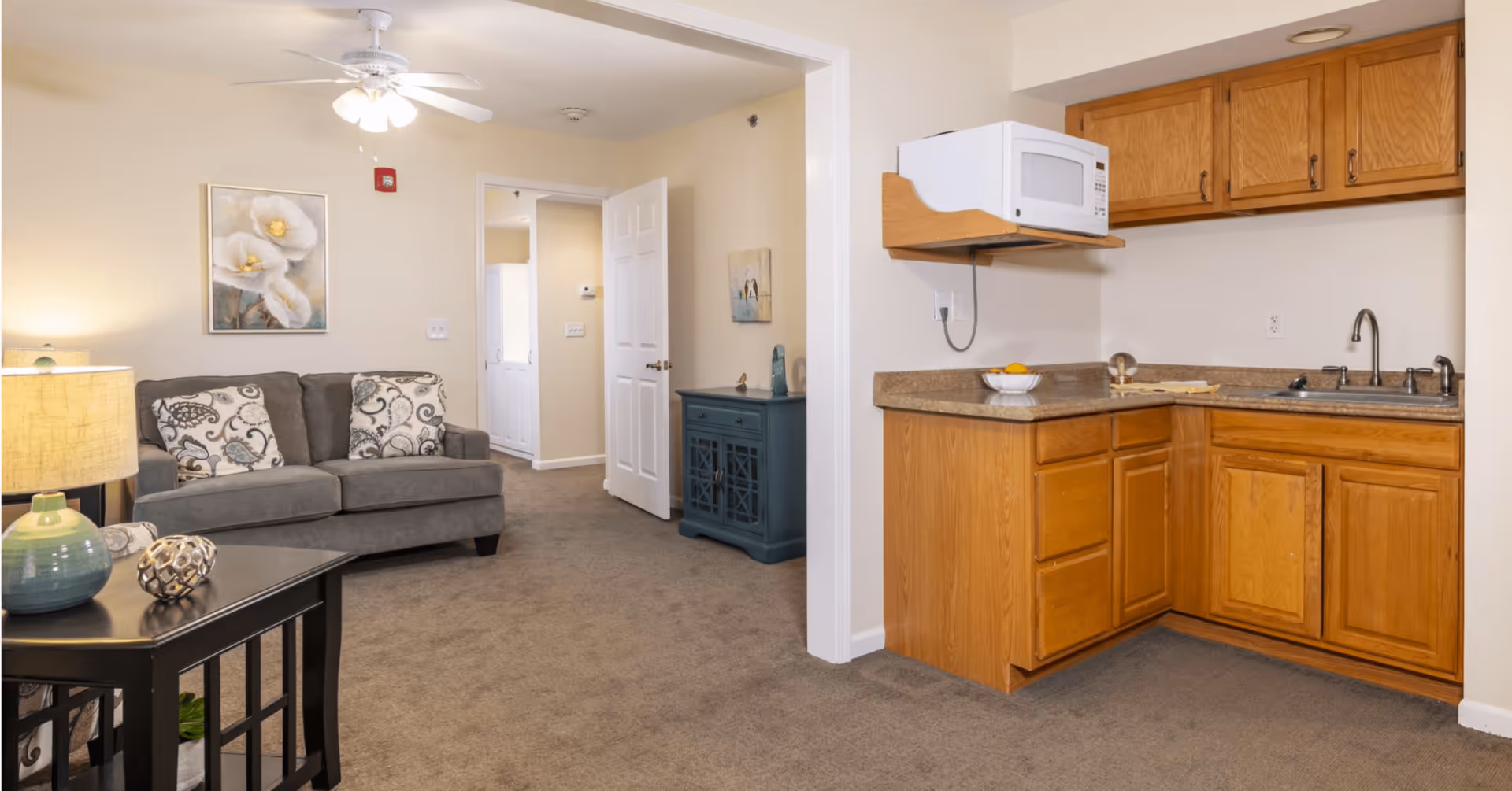 Interior view of a senior living facility showing a small kitchen area with wooden cabinets, a microwave, and a sink on the right. On the left, there is a living room area with a gray couch adorned with patterned pillows, a side table with a lamp, and a ceiling fan with lights. The walls are light-colored, and there is a doorway leading to another room in the background.