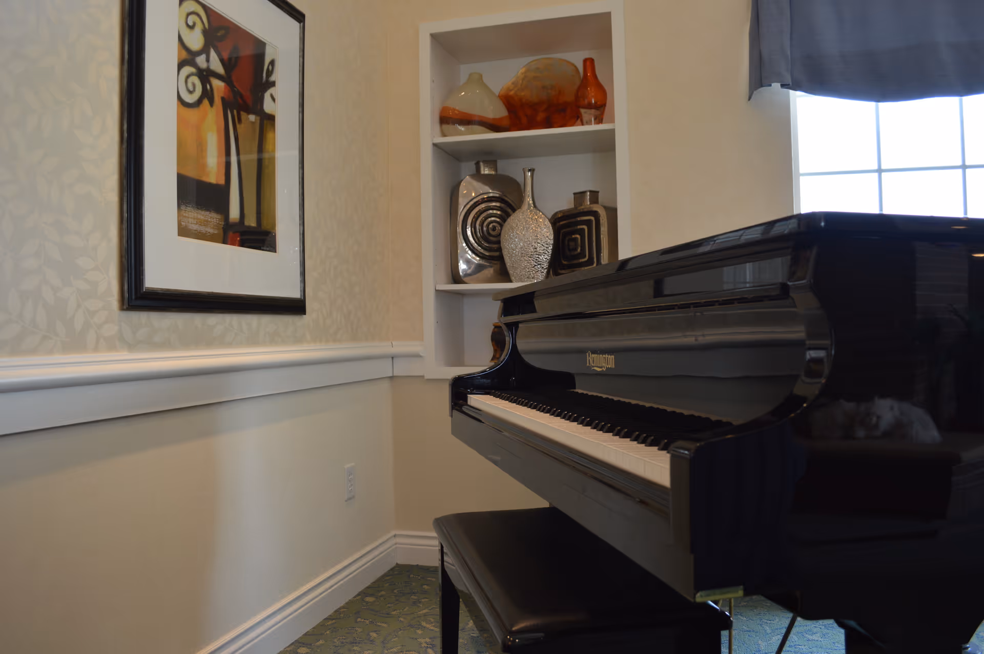 A black grand piano with a matching bench is positioned near a wall with a framed abstract painting. Built-in shelves above the piano display decorative vases. A window with a blue curtain allows natural light into the room.