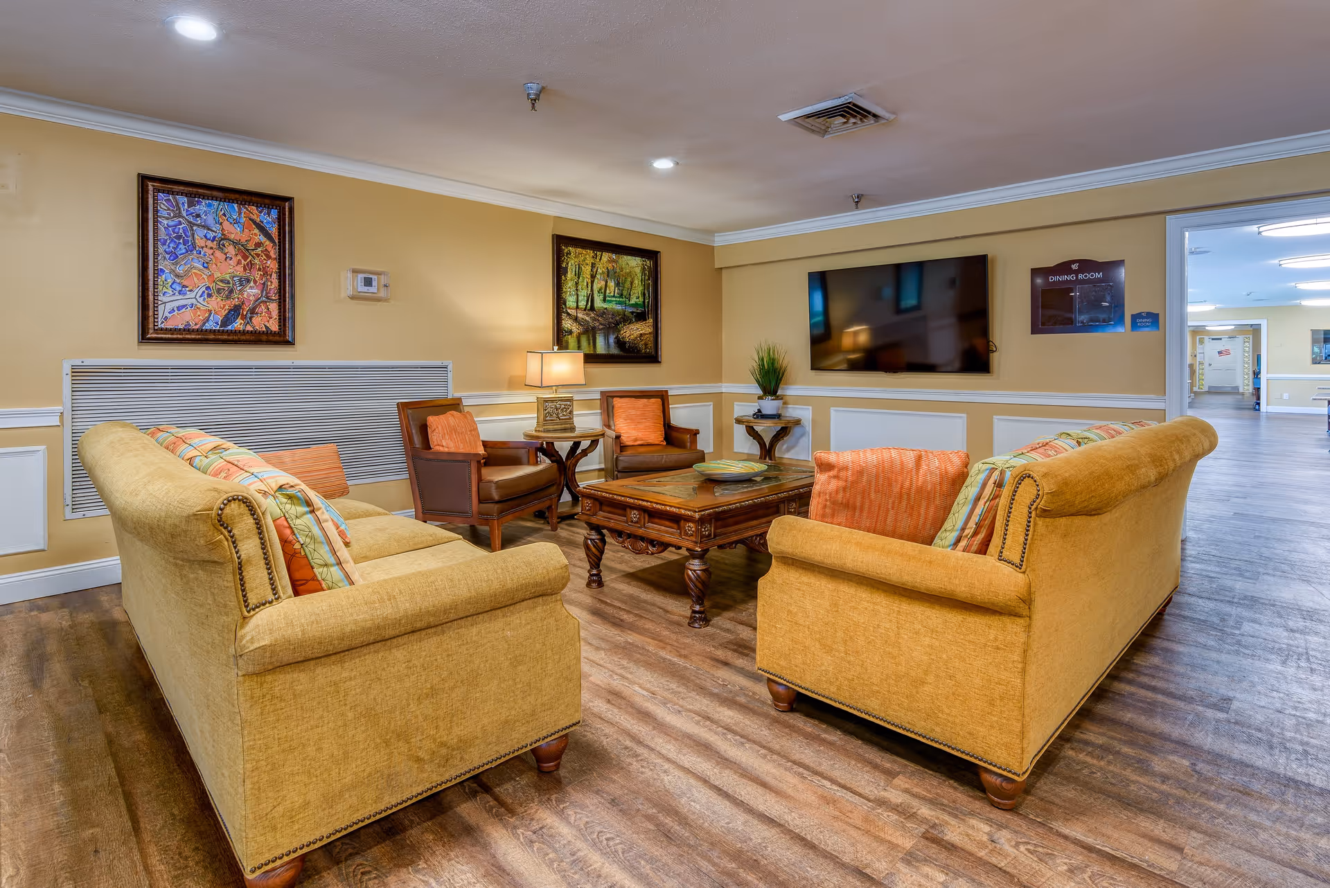 A cozy living room area with two mustard yellow sofas facing each other, two brown leather armchairs, a wooden coffee table in the center, and a flat-screen TV mounted on the wall. The walls are painted beige with white trim, and there are two framed paintings and a table lamp. The floor is wooden, and a hallway leading to a dining room is visible in the background.