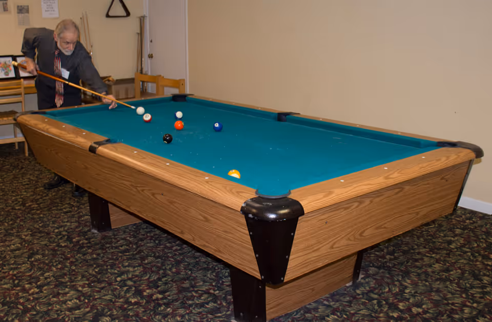 An elderly man playing pool in a room with a green pool table, several billiard balls on the table, and a cue stick in his hand. The room has patterned carpet, a wooden chair, and a beige wall with a door and a rack holding pool cues.