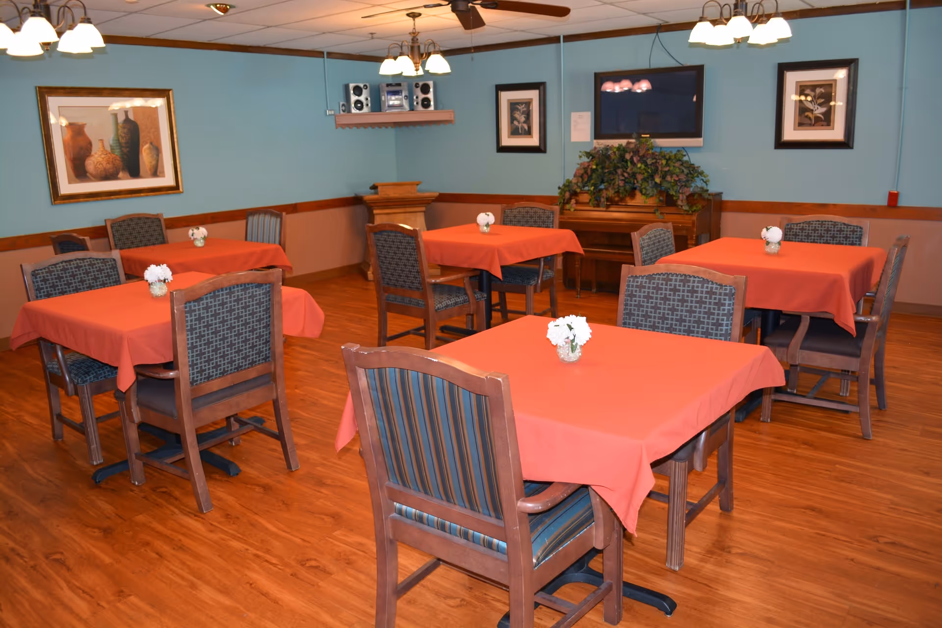 A dining room with several square tables covered with red tablecloths, each table having a small white flower centerpiece. The room has wooden flooring, blue walls with a wooden trim, framed artwork on the walls, a piano with a plant on top, and ceiling fans with lights.