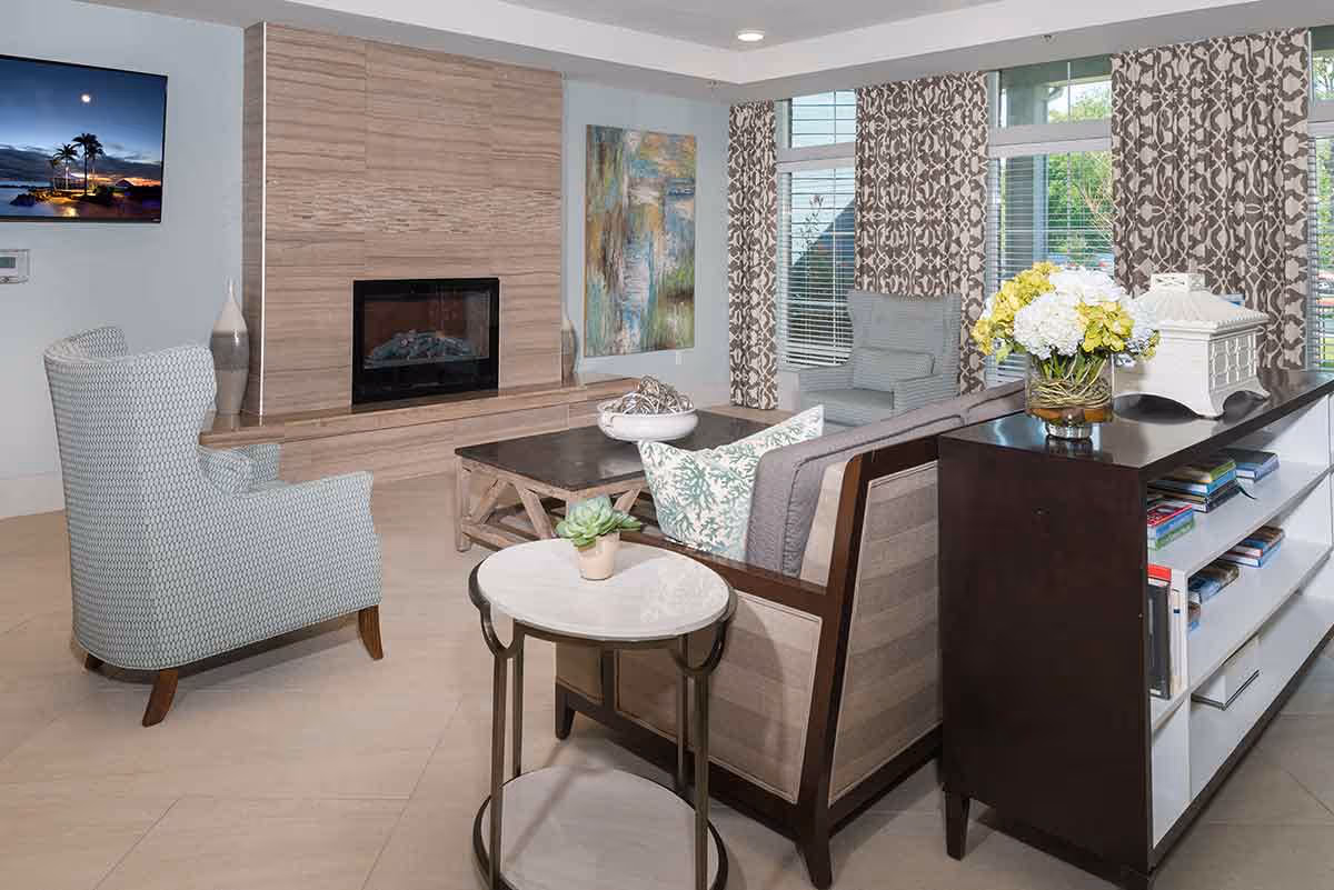 Modern communal living room with sofas and armchairs arranged around a fireplace, a wall-mounted TV, and a console table with flowers by large windows.