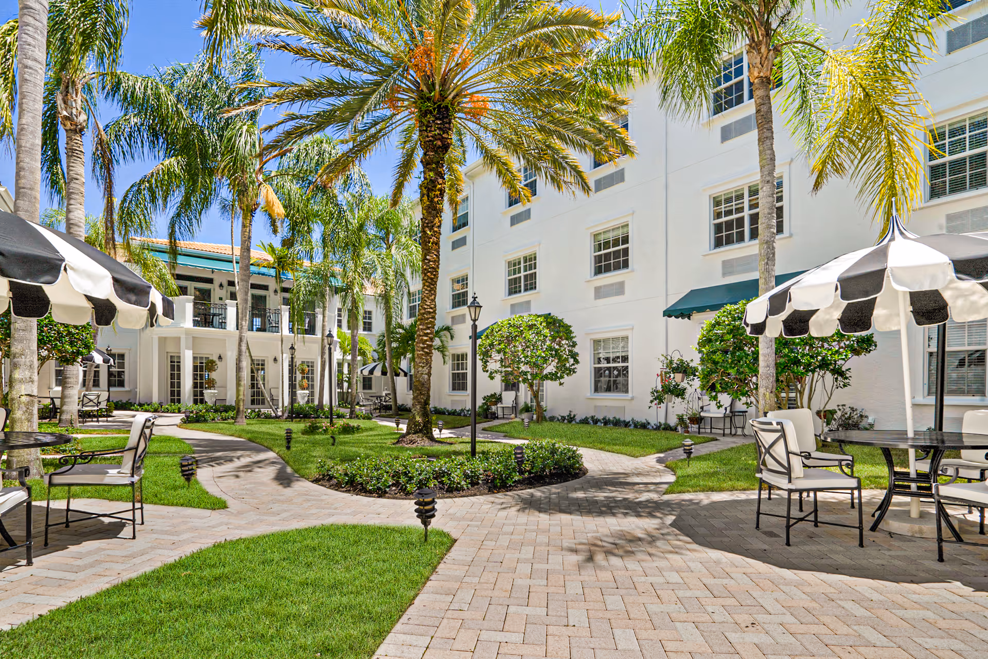 Sunny landscaped courtyard with palm trees, patio seating and black-and-white umbrellas beside a white multi-story building.