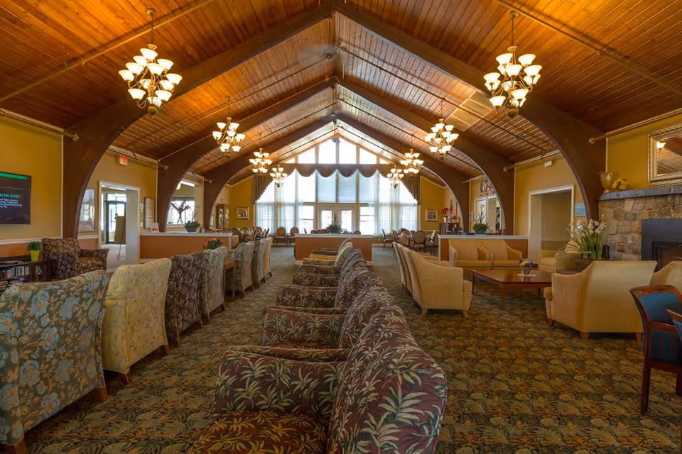 Spacious living room area in Bentley Assisted Living at Branchville with a high wooden vaulted ceiling, multiple chandeliers, patterned carpet, and rows of upholstered armchairs and sofas arranged for socializing. Large windows with sheer curtains allow natural light to fill the room. There is a stone fireplace on the right side and various plants and decorations throughout the space.