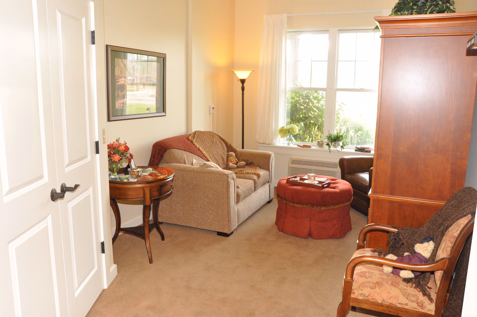 Sunlit living room with a loveseat, armchair, red ottoman, wooden armoire, side table with flowers, and a window with potted plants.
