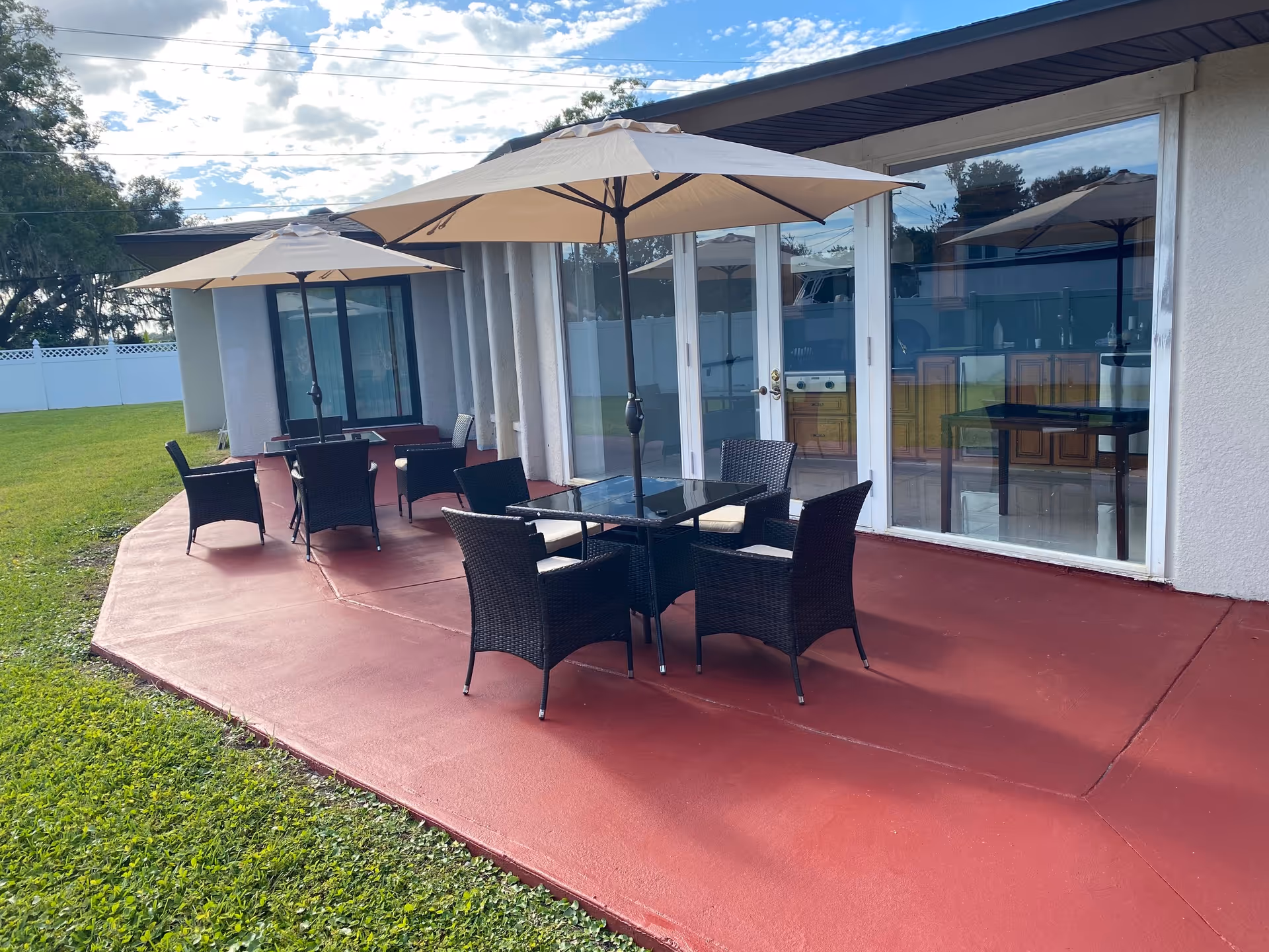 Outdoor patio area with two sets of black wicker chairs and glass tables, each shaded by a large beige umbrella. The patio has a red concrete floor and is adjacent to a building with large glass sliding doors and windows. Green grass surrounds the patio, and the sky is partly cloudy.