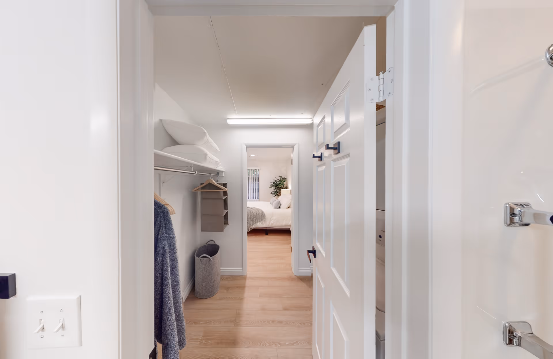 View down a hallway with a closet on the left containing hanging clothes, pillows on a shelf, and a laundry basket on the floor. At the end of the hallway, a bedroom with a bed, pillows, and a plant near a window is visible. The right side shows part of a white door and a glimpse of a bathroom with a shower.