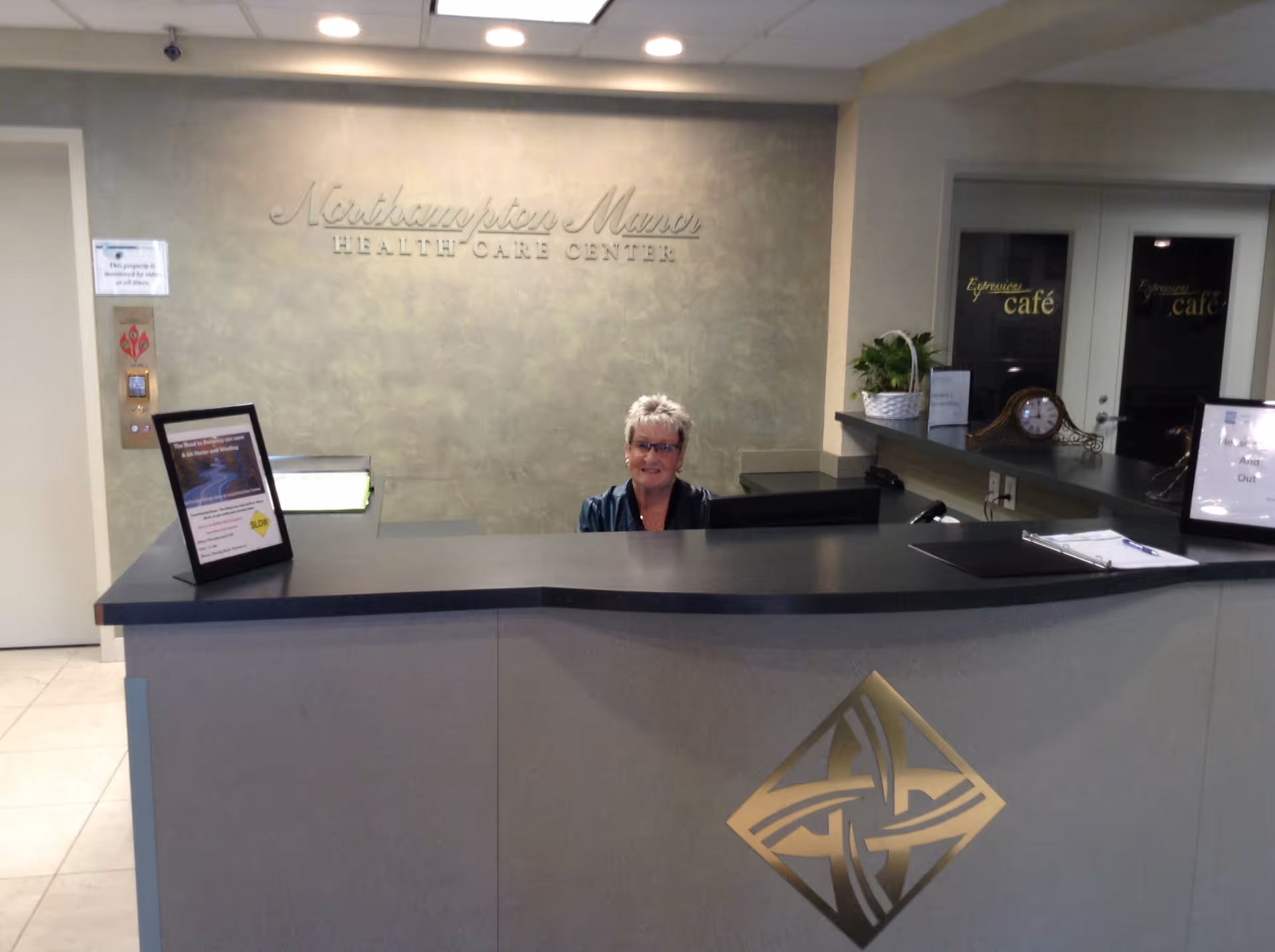 Reception desk area at Northampton Manor Health Care Center with a smiling receptionist behind the counter. The wall behind has the facility name displayed, and there are signs and a clock on the counter. The entrance to the Espresso cafe is visible in the background.